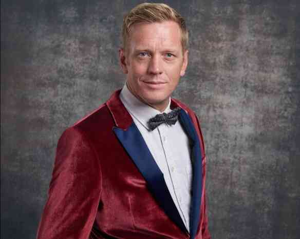 A man with short blonde hair is wearing a velvet burgundy suit jacket with a blue lapel and a white shirt underneath. He is also sporting a black bow tie and is smiling confidently, set against a textured gray background.