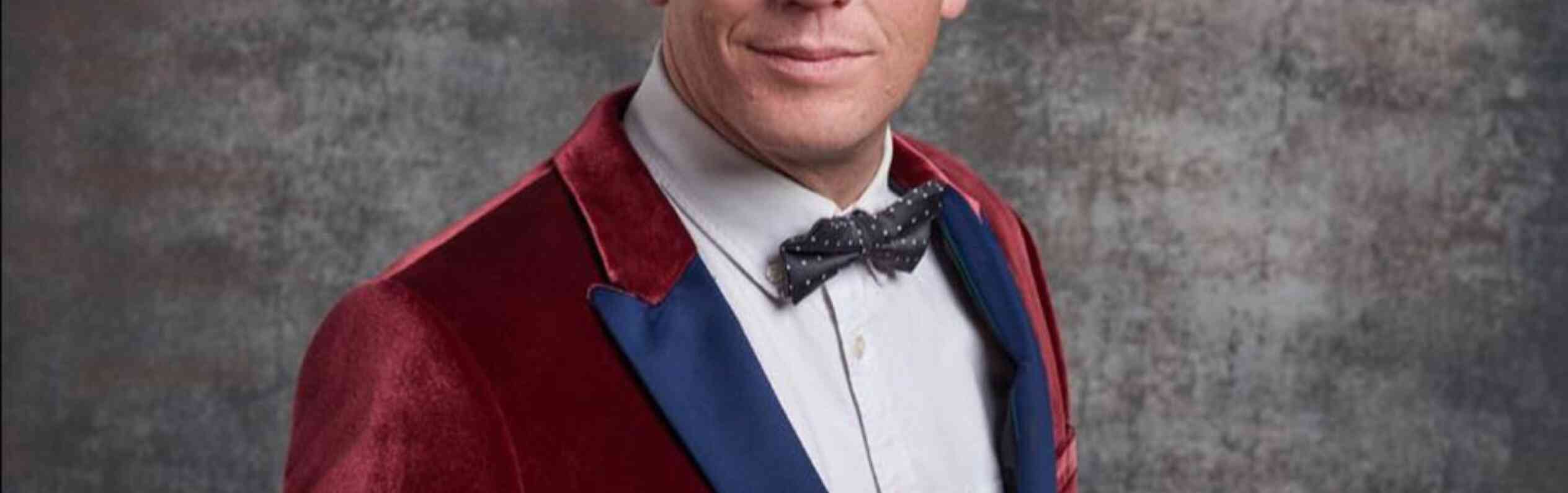 A man with short blonde hair is wearing a velvet burgundy suit jacket with a blue lapel and a white shirt underneath. He is also sporting a black bow tie and is smiling confidently, set against a textured gray background.