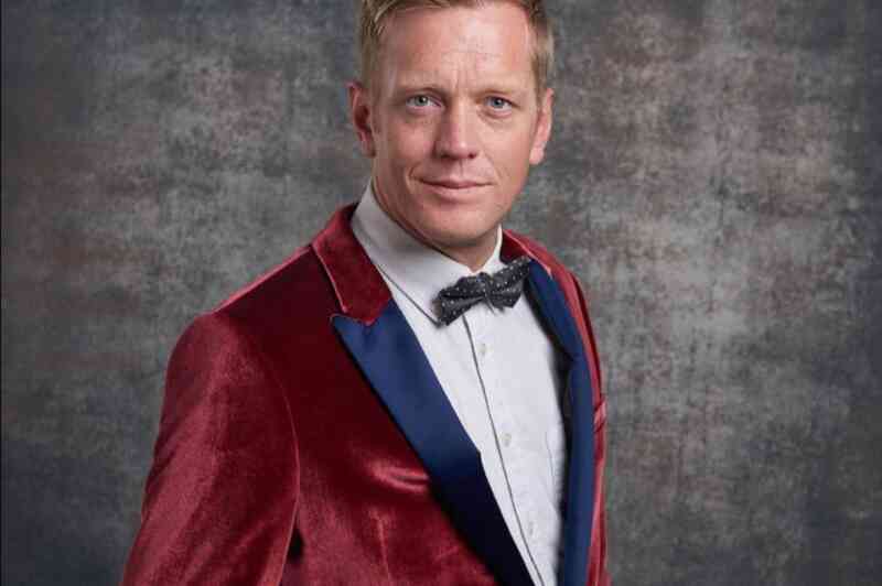 A man with short blonde hair is wearing a velvet burgundy suit jacket with a blue lapel and a white shirt underneath. He is also sporting a black bow tie and is smiling confidently, set against a textured gray background.