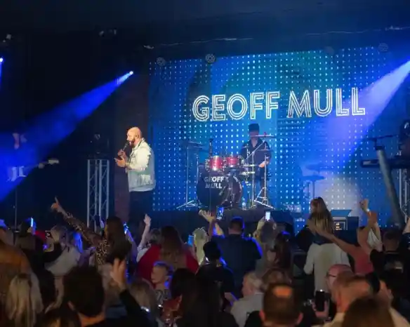 A lively concert scene with a singer performing on stage in front of a large audience. A drummer is visible in the background, and bright blue lights and a digital display showcase the name "Geoff Mull." The crowd appears engaged and excited.