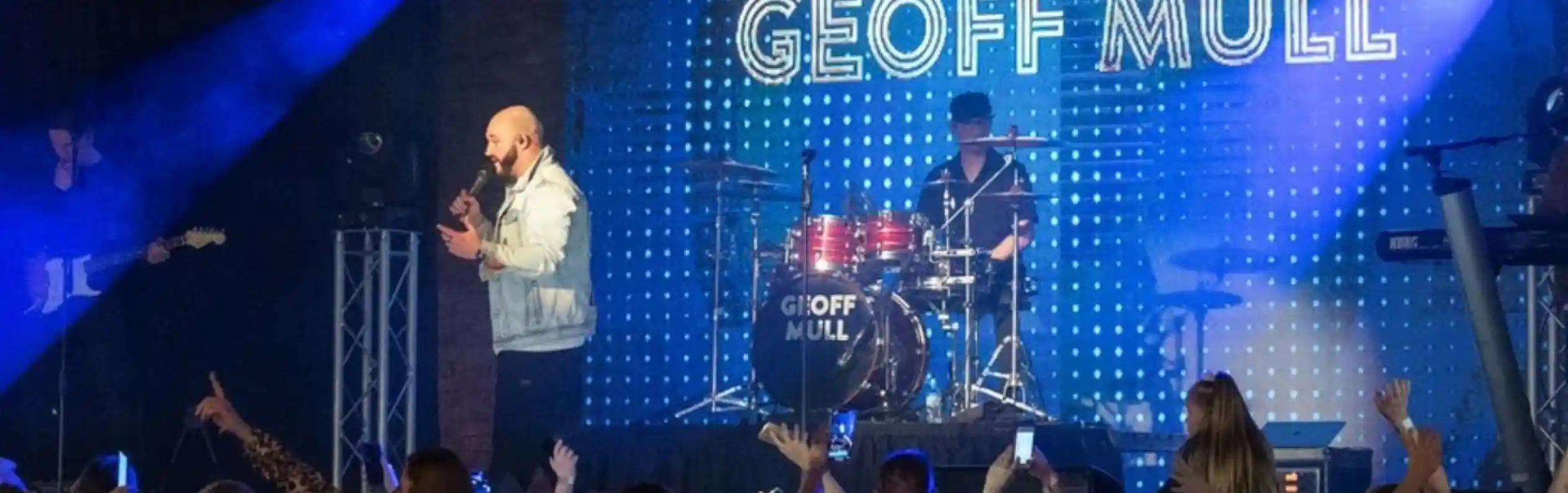 A lively concert scene with a singer performing on stage in front of a large audience. A drummer is visible in the background, and bright blue lights and a digital display showcase the name "Geoff Mull." The crowd appears engaged and excited.