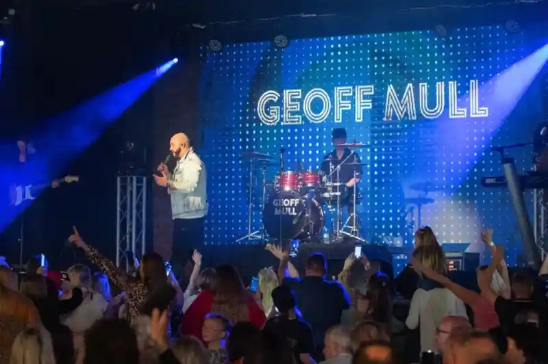 A lively concert scene with a singer performing on stage in front of a large audience. A drummer is visible in the background, and bright blue lights and a digital display showcase the name "Geoff Mull." The crowd appears engaged and excited.