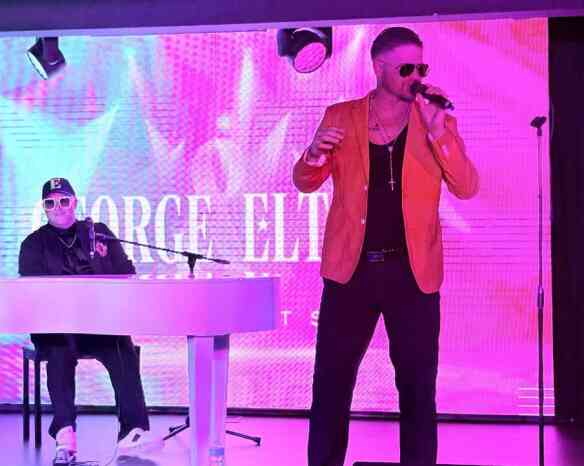 A performer in a bright orange jacket sings into a microphone, while a musician in a black outfit plays a white keyboard. The backdrop features vibrant, colorful visuals.