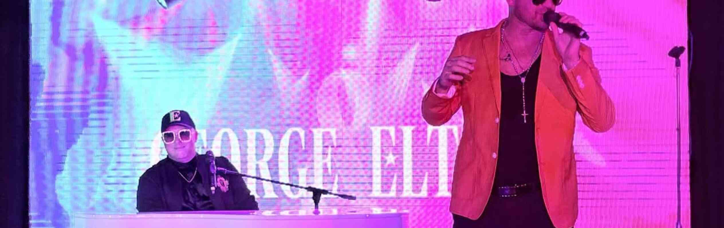 A performer in a bright orange jacket sings into a microphone, while a musician in a black outfit plays a white keyboard. The backdrop features vibrant, colorful visuals.