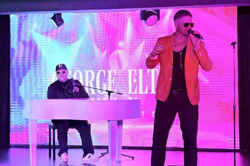 A performer in a bright orange jacket sings into a microphone, while a musician in a black outfit plays a white keyboard. The backdrop features vibrant, colorful visuals.