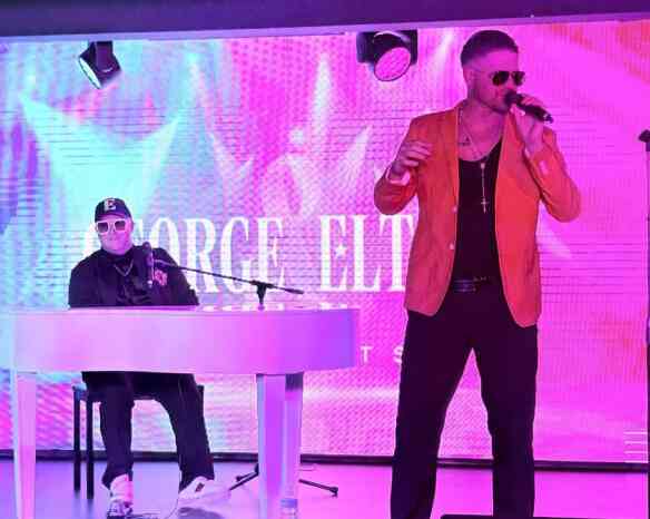 A performer in a bright orange jacket sings into a microphone, while a musician in a black outfit plays a white keyboard. The backdrop features vibrant, colorful visuals.