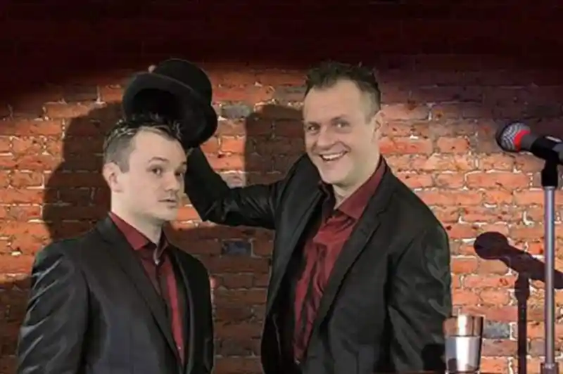 Two men in black suits stand in front of a brick wall. One is holding a top hat while smiling, and the other looks surprised. A microphone and a stool with a glass of water are positioned nearby.