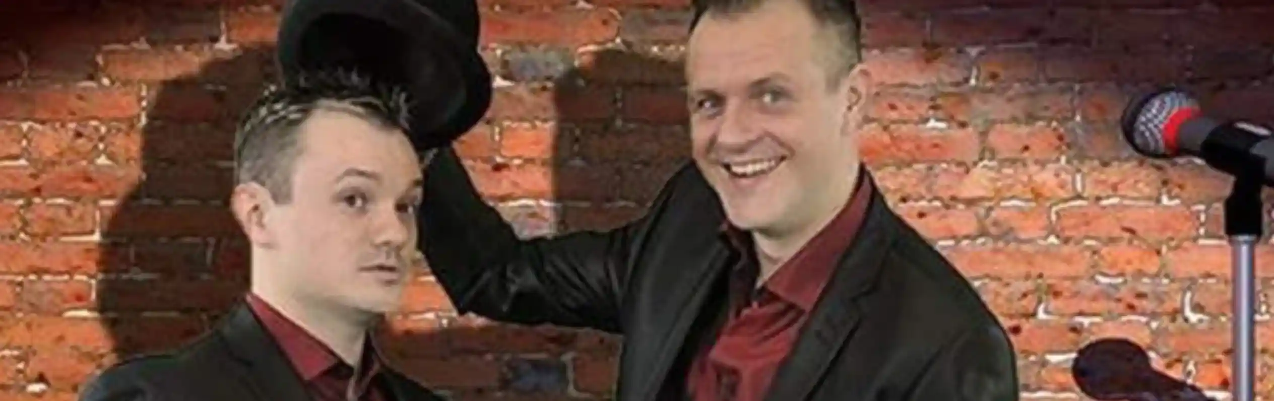 Two men in black suits stand in front of a brick wall. One is holding a top hat while smiling, and the other looks surprised. A microphone and a stool with a glass of water are positioned nearby.