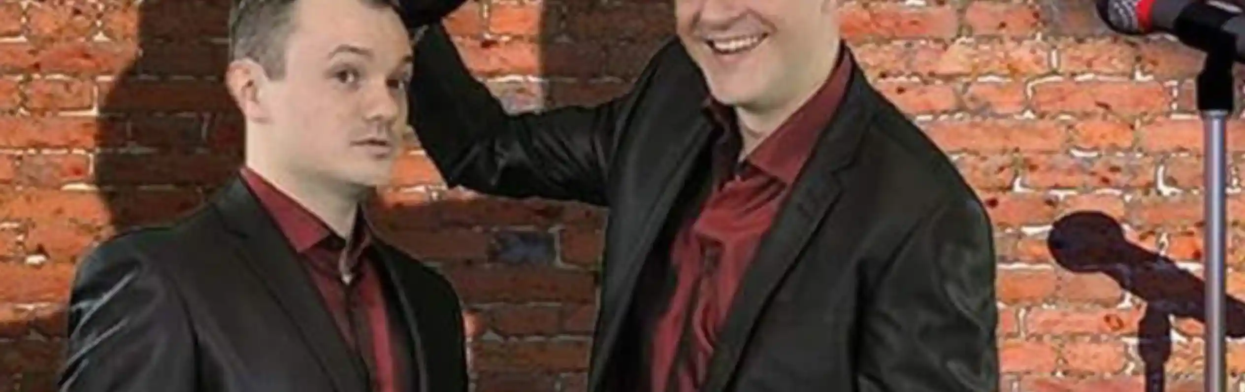 Two men in black suits stand in front of a brick wall. One is holding a top hat while smiling, and the other looks surprised. A microphone and a stool with a glass of water are positioned nearby.