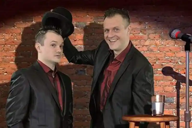 Two men in black suits stand in front of a brick wall. One is holding a top hat while smiling, and the other looks surprised. A microphone and a stool with a glass of water are positioned nearby.