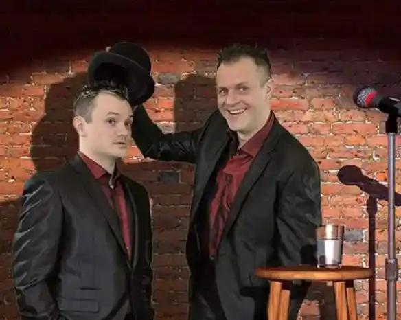 Two men in black suits stand in front of a brick wall. One is holding a top hat while smiling, and the other looks surprised. A microphone and a stool with a glass of water are positioned nearby.