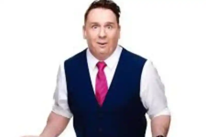 A man in a blue vest and white shirt with a pink tie stands with an expressive face, hands raised and open, conveying surprise or confusion.
