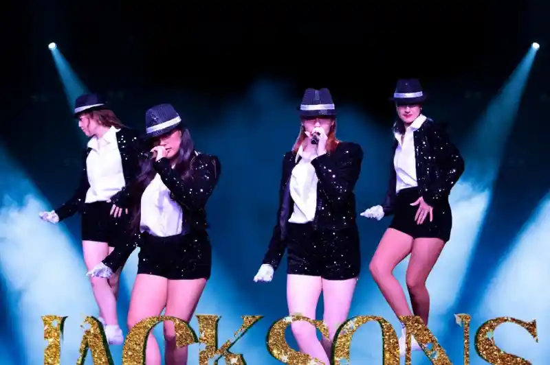 Four performers in sparkling black outfits and hats sing on stage. They are surrounded by blue lights and glitter, with the word "JACKSONS" in gold letters at the bottom.