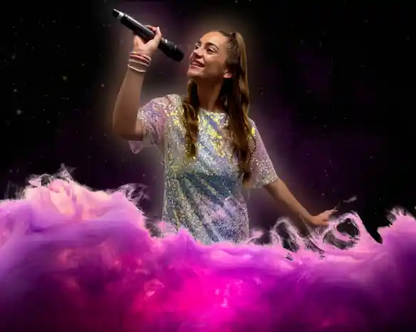 A young woman with long brown hair, wearing a sparkly silver dress, sings passionately into a microphone against a swirling pink and purple backdrop.