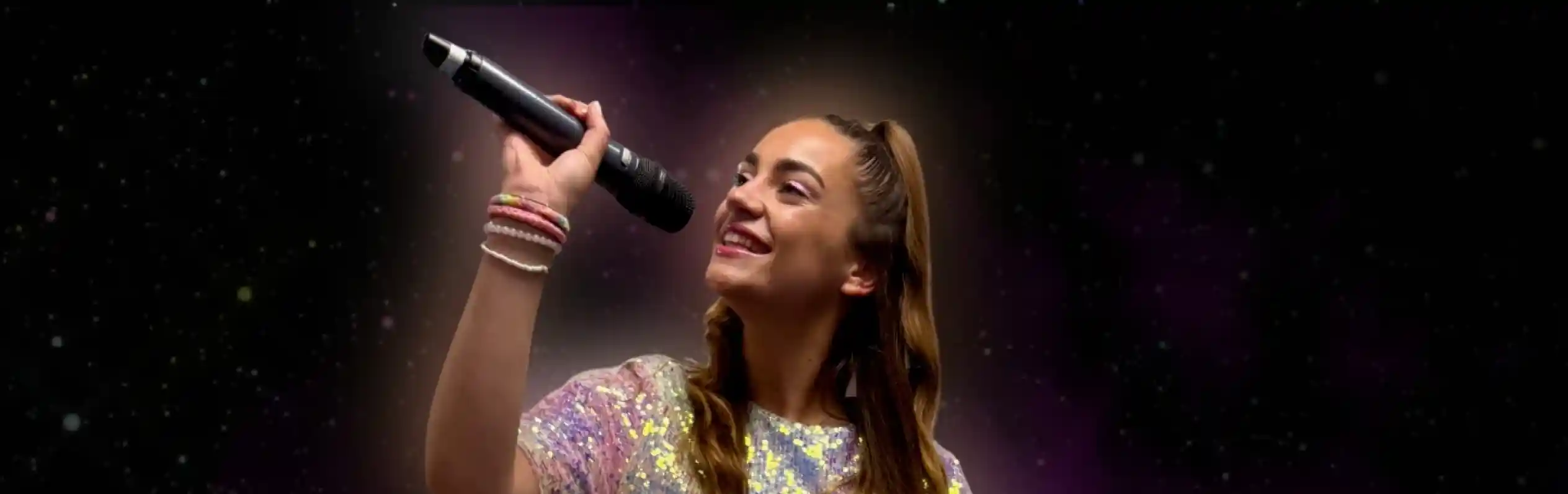 A young woman with long brown hair, wearing a sparkly silver dress, sings passionately into a microphone against a swirling pink and purple backdrop.