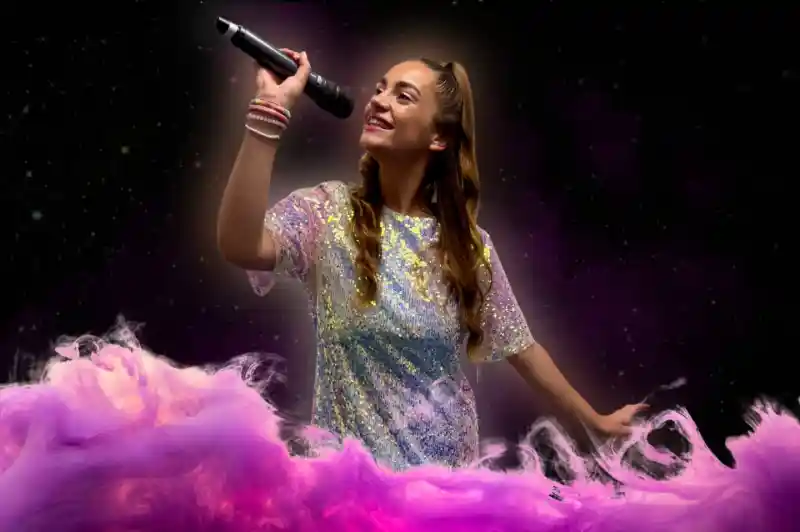 A young woman with long brown hair, wearing a sparkly silver dress, sings passionately into a microphone against a swirling pink and purple backdrop.