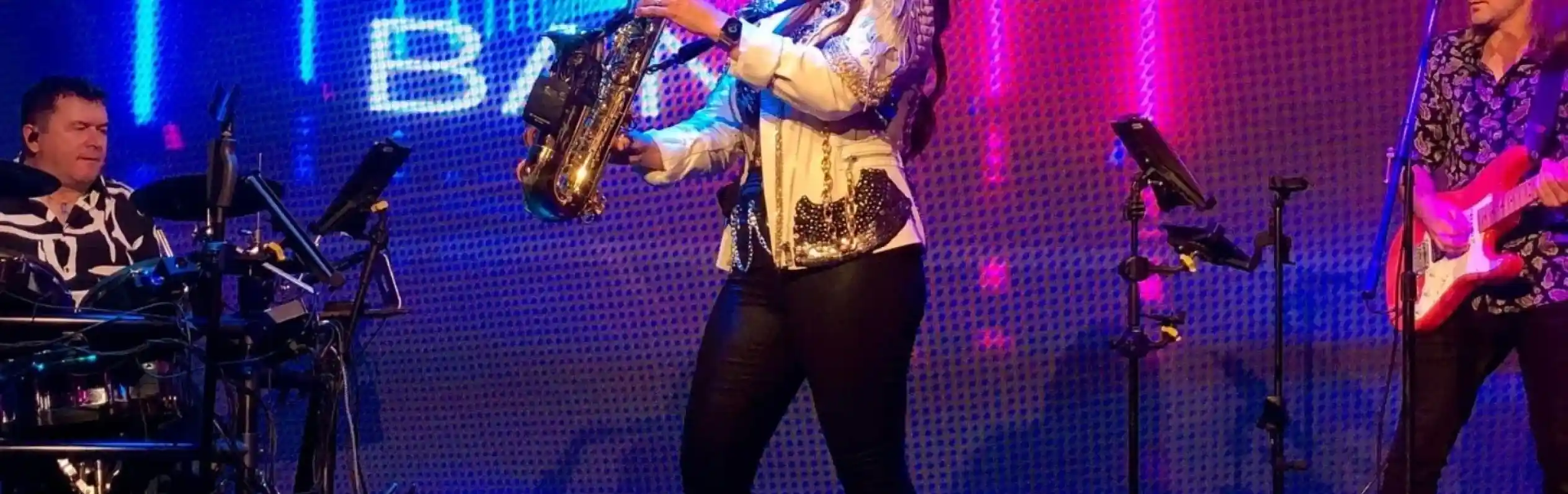 A female saxophonist performs on stage in fashionable attire, holding a saxophone. She is flanked by a drummer on the left and a guitarist on the right, both engaged in the performance. Colorful lights and a neon "Jet Set Band" logo illuminate the background.