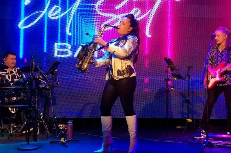 A female saxophonist performs on stage in fashionable attire, holding a saxophone. She is flanked by a drummer on the left and a guitarist on the right, both engaged in the performance. Colorful lights and a neon "Jet Set Band" logo illuminate the background.