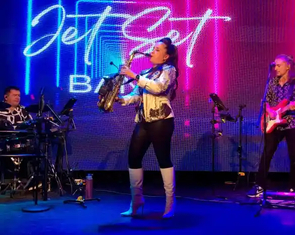 A female saxophonist performs on stage in fashionable attire, holding a saxophone. She is flanked by a drummer on the left and a guitarist on the right, both engaged in the performance. Colorful lights and a neon "Jet Set Band" logo illuminate the background.