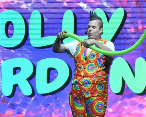 A performer in a colorful polka-dotted outfit creates a balloon sculpture on stage, with bright, playful graphics in the background.