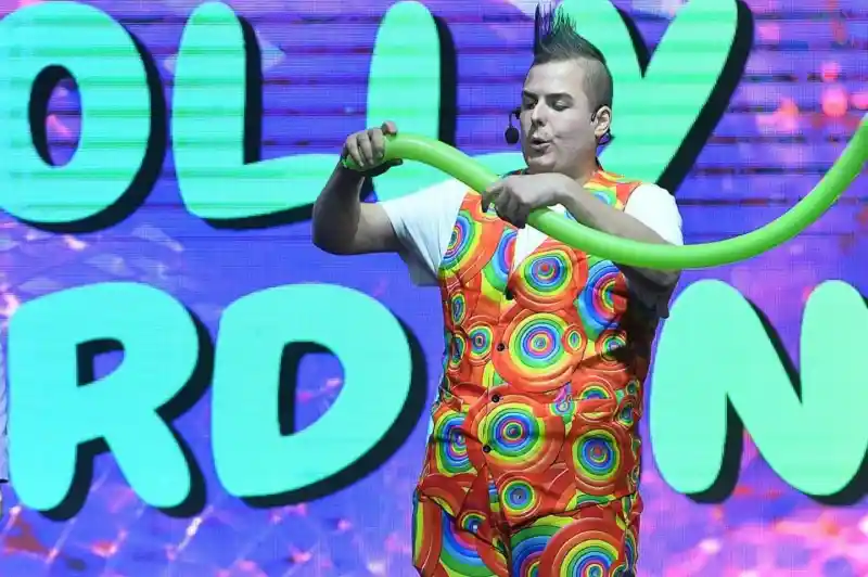 A performer in a colorful polka-dotted outfit creates a balloon sculpture on stage, with bright, playful graphics in the background.
