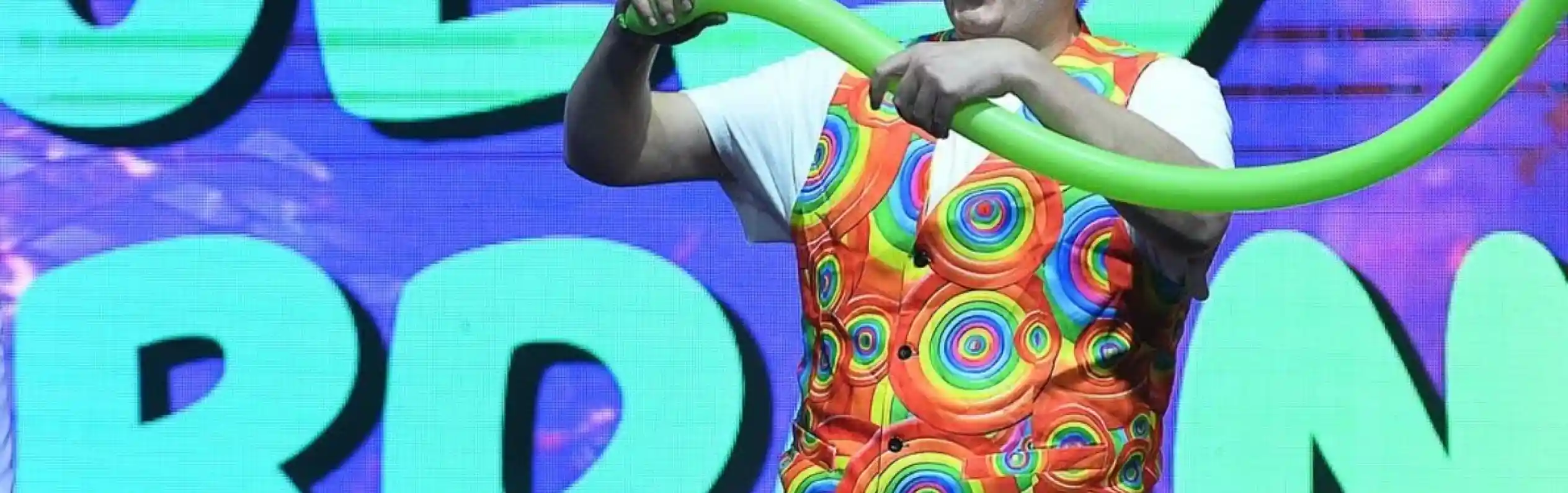 A performer in a colorful polka-dotted outfit creates a balloon sculpture on stage, with bright, playful graphics in the background.