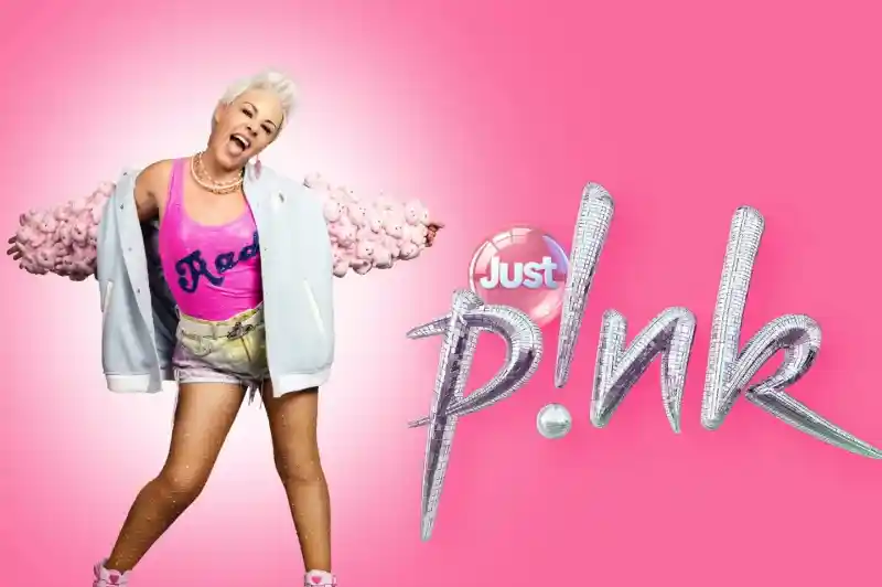 A person with short blonde hair is posing energetically in a pink-themed outfit, featuring a bright pink tank top and a light blue jacket with fluffy pink accents. They have a playful expression and are wearing pink high-top sneakers, set against a vibrant pink background with the stylized text "Just p!nk."