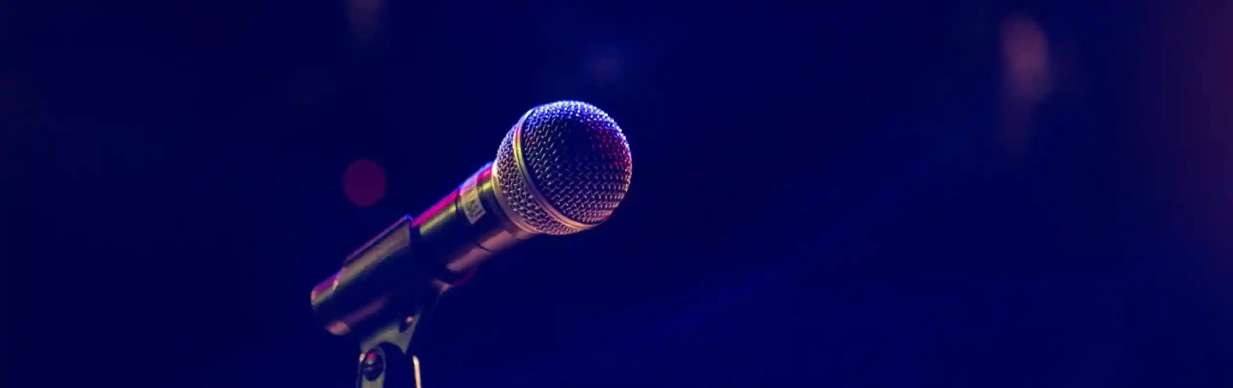 A close-up of a microphone standing on a stage, with a blurred background featuring dim lighting and hints of colorful lights.