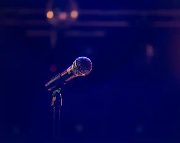 A close-up of a microphone standing on a stage, with a blurred background featuring dim lighting and hints of colorful lights.