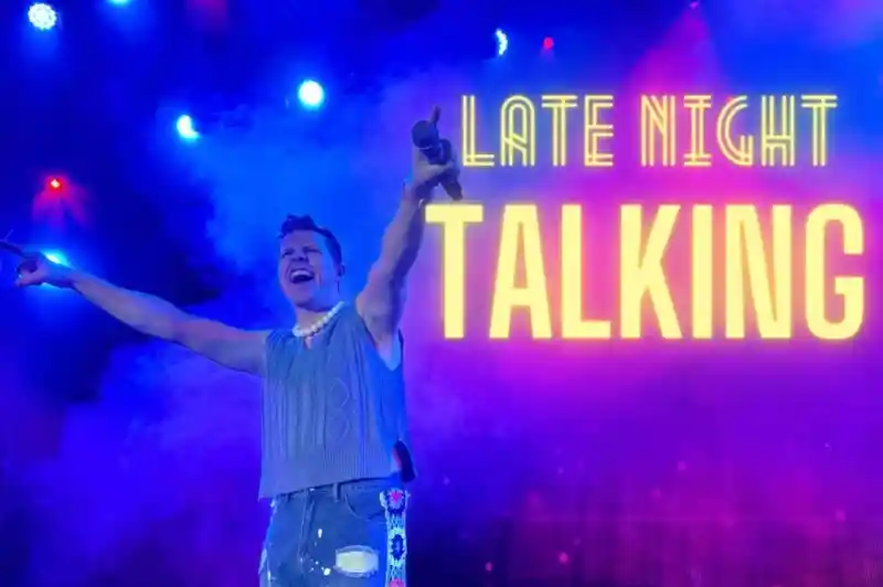 A performer with a joyful expression stands on stage, holding a microphone and wearing a sleeveless sweater and embellished jeans. Colorful lights and smoke create a vibrant atmosphere, with "Late Night Talking" prominently displayed in bold text.