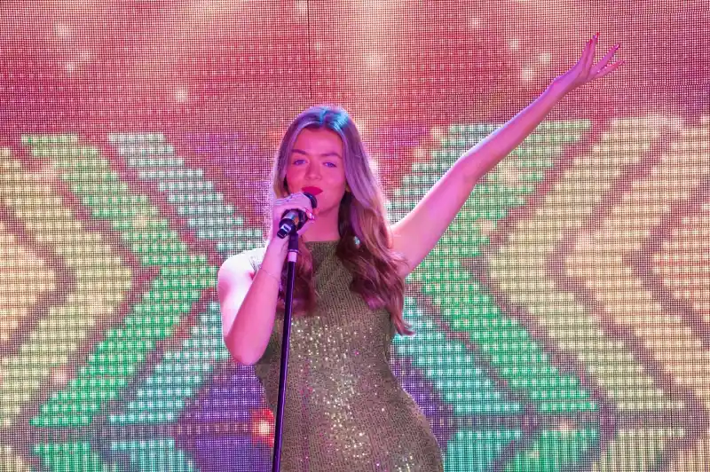 A lady is singing on stage into a microphone. She is wearing a sparkly gold dress and has one arm up in the air posing against a sparkly screen backdrop.