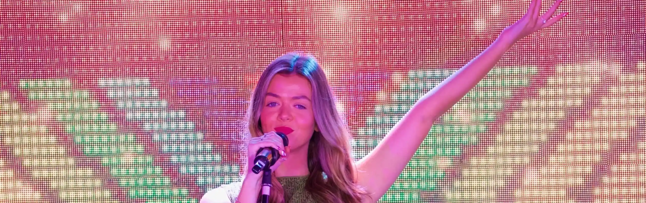 A lady is singing on stage into a microphone. She is wearing a sparkly gold dress and has one arm up in the air posing against a sparkly screen backdrop.