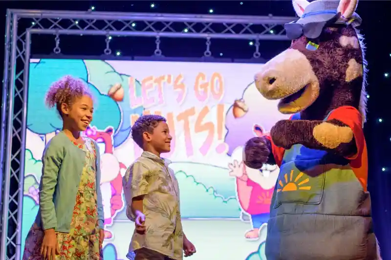 A costumed performer stands on stage with two young children who are smiling and laughing with the performer, with a screen in the background with the words 'Let's Go Nuts!'