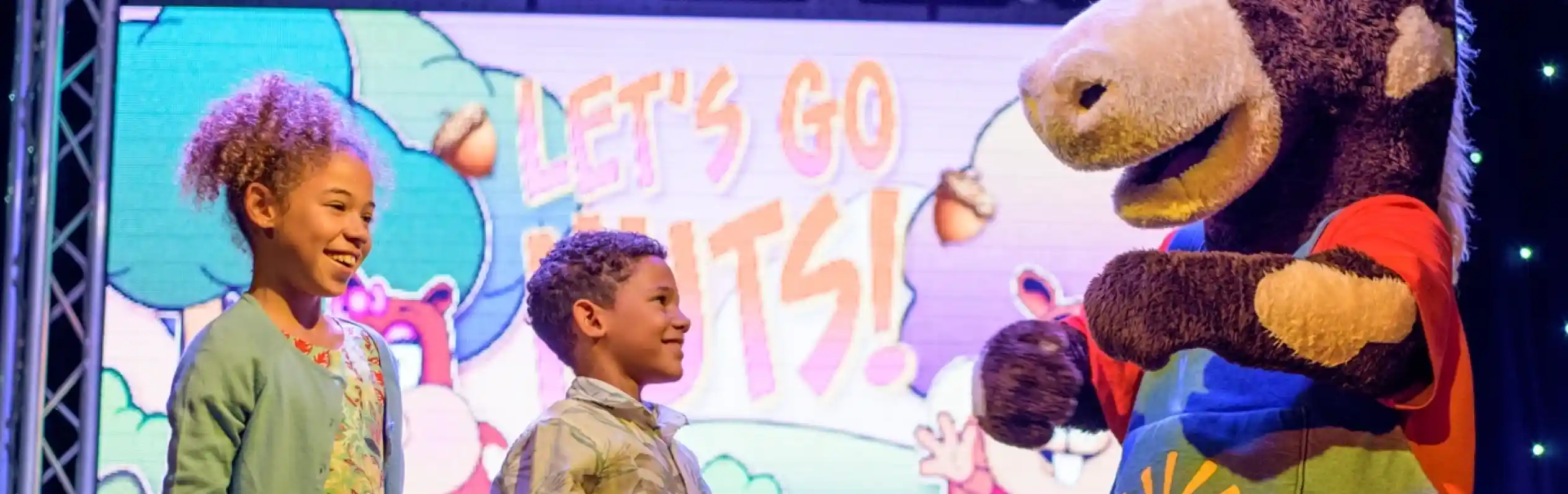 A costumed performer stands on stage with two young children who are smiling and laughing with the performer, with a screen in the background with the words 'Let's Go Nuts!'