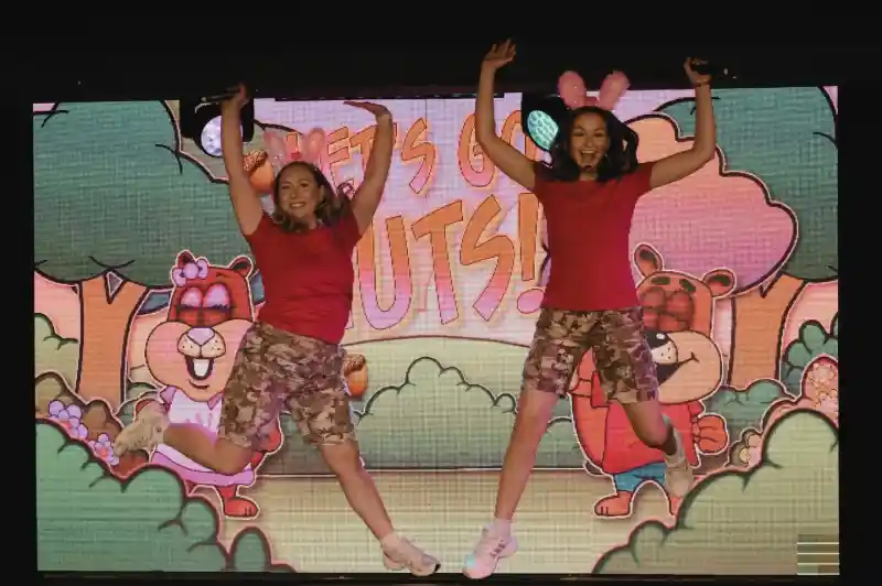 Two women in pink shirts and camo shorts perform on stage, smiling and engaging with the audience. A colorful backdrop displays the text "Let's Go Nuts!" with cartoonish trees and acorns.