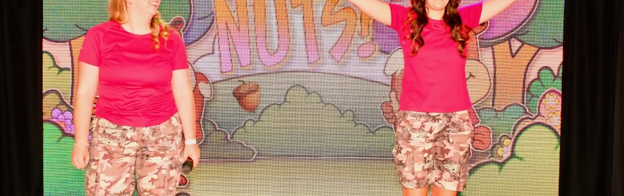 Two women in pink shirts and camo shorts perform on stage, smiling and engaging with the audience. A colorful backdrop displays the text "Let's Go Nuts!" with cartoonish trees and acorns.