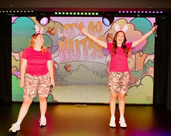 Two women in pink shirts and camo shorts perform on stage, smiling and engaging with the audience. A colorful backdrop displays the text "Let's Go Nuts!" with cartoonish trees and acorns.