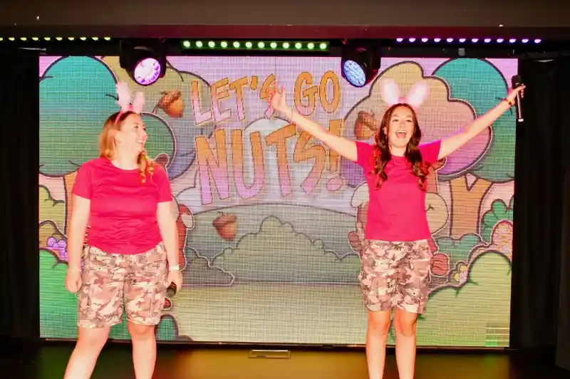 Two women in pink shirts and camo shorts perform on stage, smiling and engaging with the audience. A colorful backdrop displays the text "Let's Go Nuts!" with cartoonish trees and acorns.