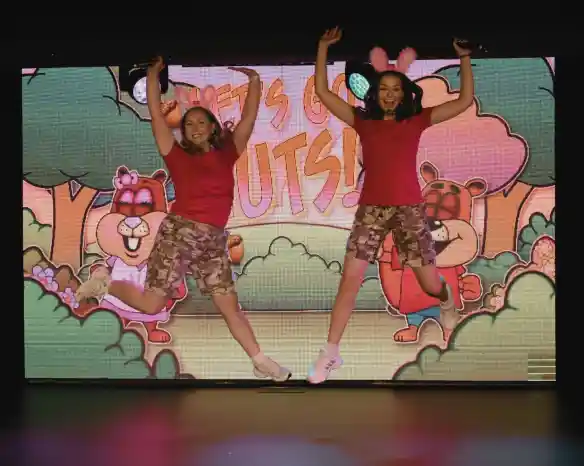 Two women in pink shirts and camo shorts perform on stage, smiling and engaging with the audience. A colorful backdrop displays the text "Let's Go Nuts!" with cartoonish trees and acorns.