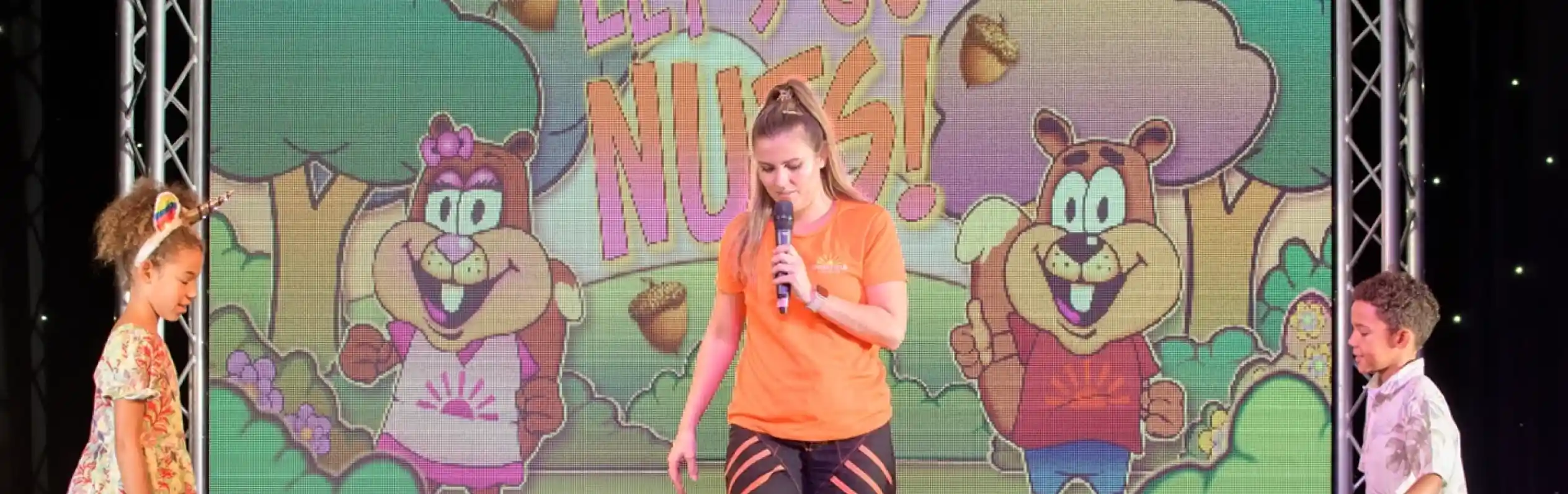 A performer on stage holds a microphone and is giving instructions to two children who are entering the stage to take part in an activity. There is a box of something unknown in the middle of the stage which will be part of the activity and a screen behind the performer projecting an image of the Squirrel Squad characters with the words Let's Go Nuts!