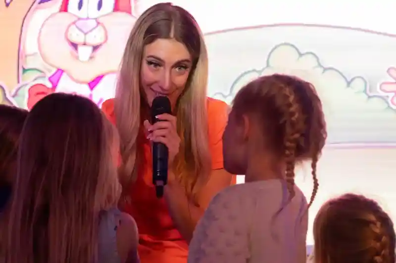 A woman with long blonde hair speaks into a microphone, engaging with a group of young children during an event. A colorful cartoon backdrop features a rabbit character.