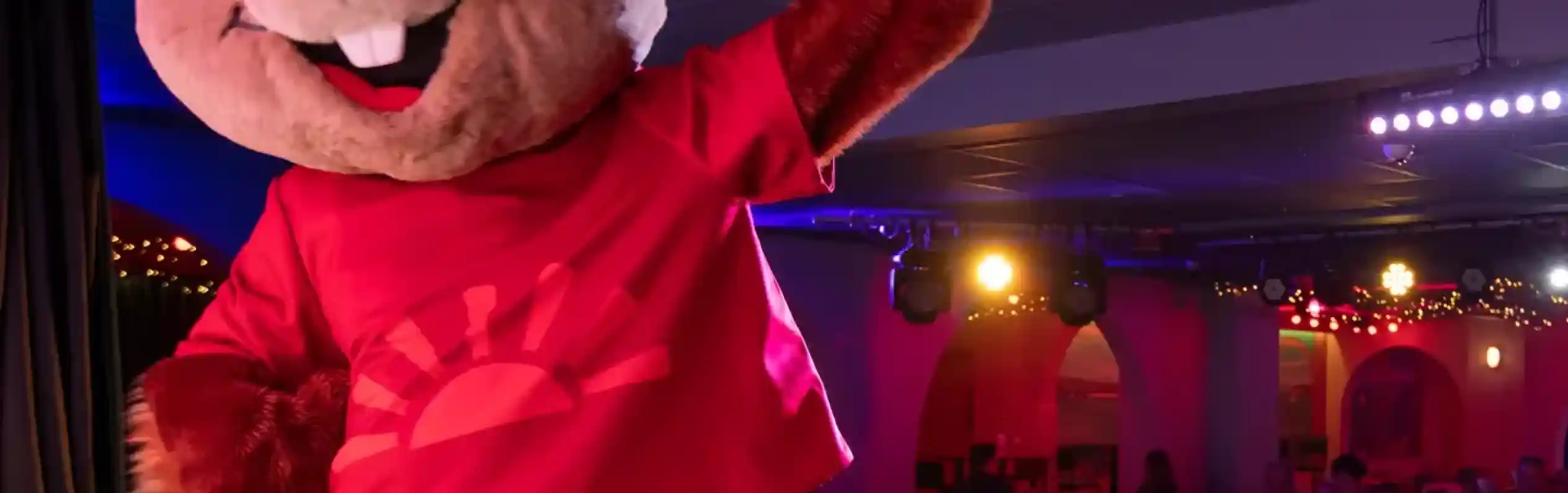 A large, smiling bear mascot in a red shirt and blue shorts energetically poses on stage, while a group of children is gathered in front, enjoying the performance. Colorful stage lights create a festive atmosphere.