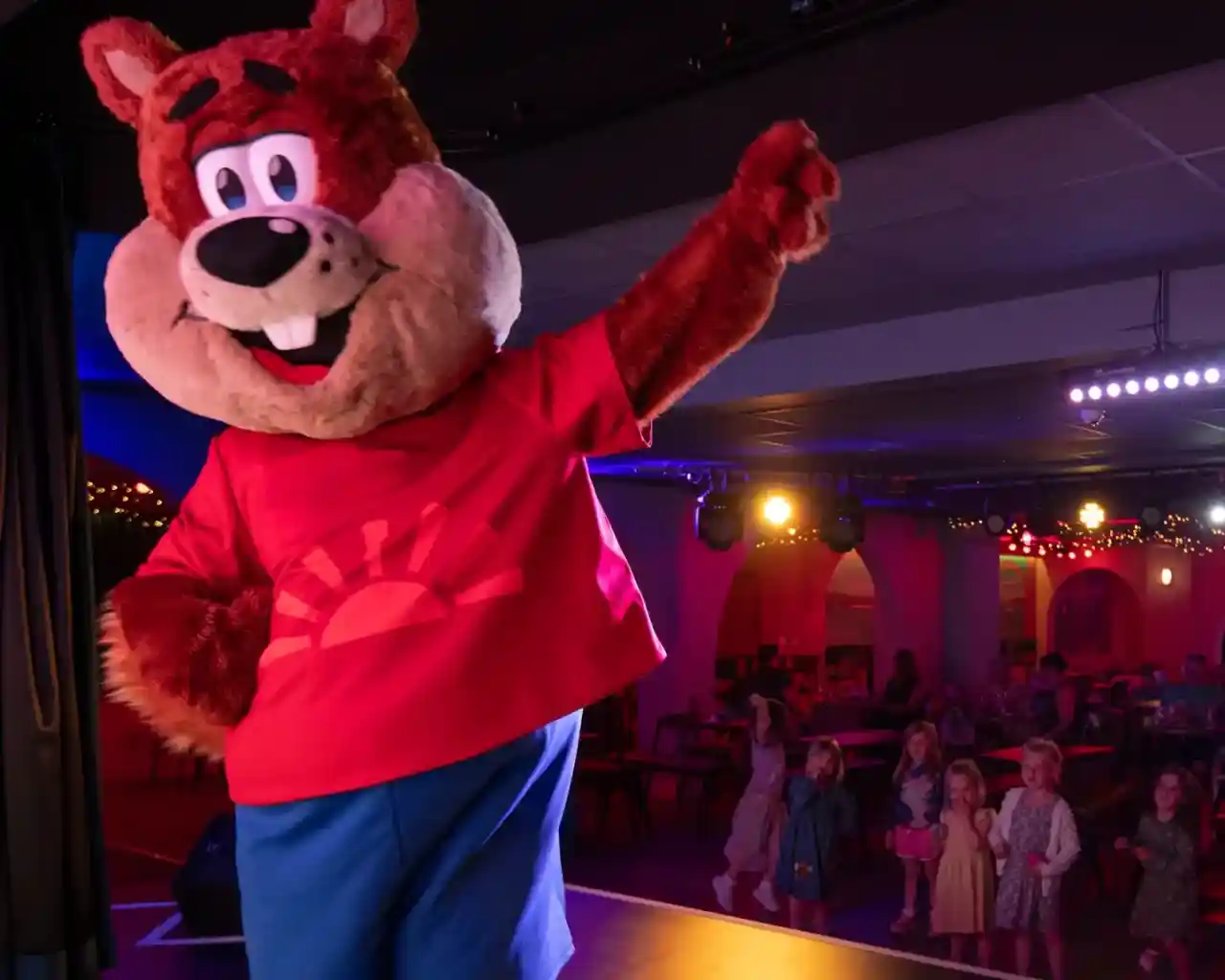 A large, smiling bear mascot in a red shirt and blue shorts energetically poses on stage, while a group of children is gathered in front, enjoying the performance. Colorful stage lights create a festive atmosphere.