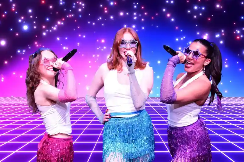 Three performers in shiny, colorful outfits with sparkly skirts and star-shaped sunglasses sing into microphones. They stand against a vibrant, starry background with a grid pattern. The scene has a fun, retro vibe, reminiscent of a disco party.