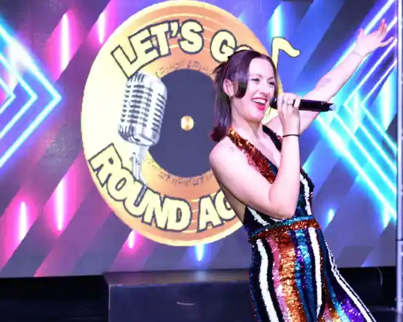 A female singer in a glittering, multicolored dress performs energetically on stage, holding a microphone. Behind her is a colorful backdrop featuring the text "Let's Go Round Again" and a microphone graphic.
