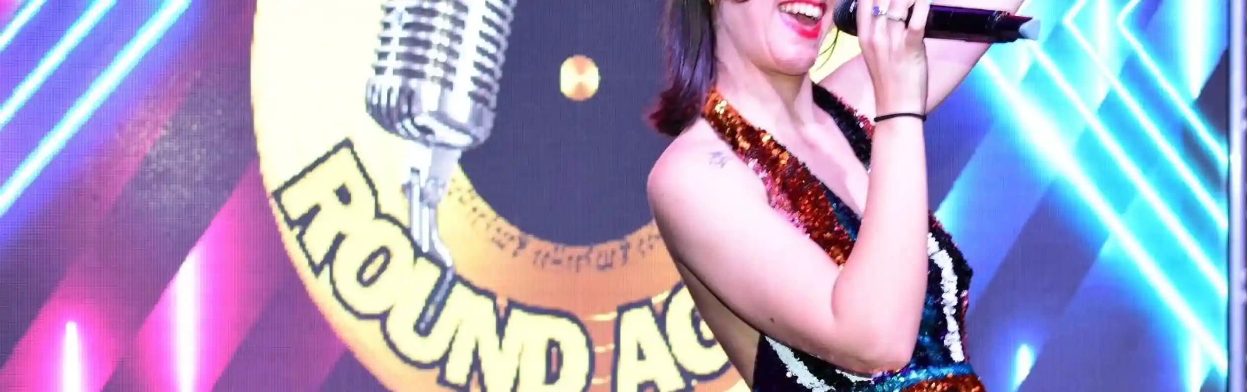 A female singer in a glittering, multicolored dress performs energetically on stage, holding a microphone. Behind her is a colorful backdrop featuring the text "Let's Go Round Again" and a microphone graphic.