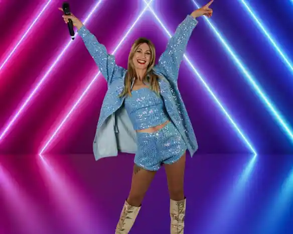 A woman poses joyfully with arms raised, wearing a sparkly blue outfit and knee-high boots, standing in front of vibrant neon lights in pink and blue hues. She holds a microphone and has a big smile on her face.