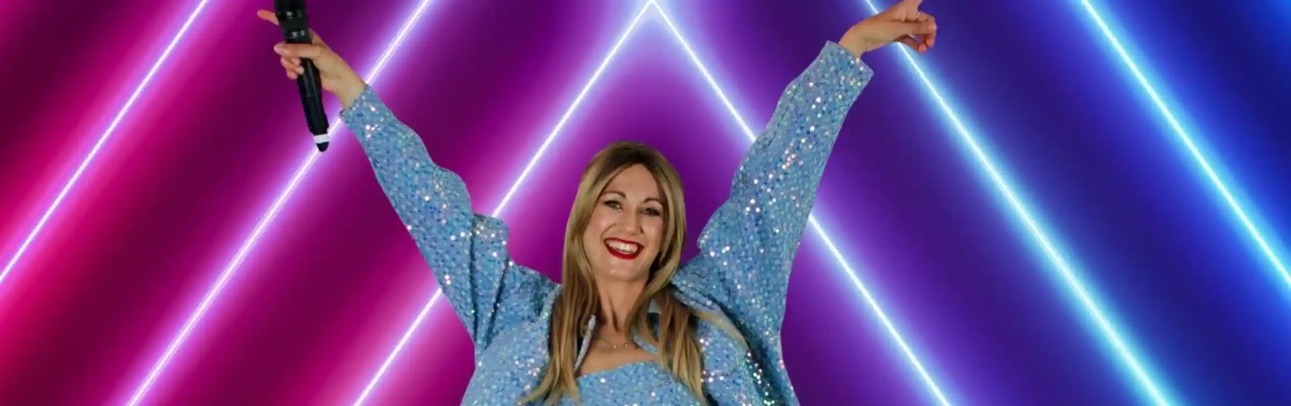 A woman poses joyfully with arms raised, wearing a sparkly blue outfit and knee-high boots, standing in front of vibrant neon lights in pink and blue hues. She holds a microphone and has a big smile on her face.