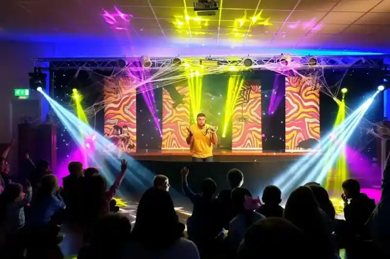 A performer stands on stage with colorful lights and projections behind them, entertaining an engaged audience seated on the floor. Bright beams of light create a dynamic atmosphere.