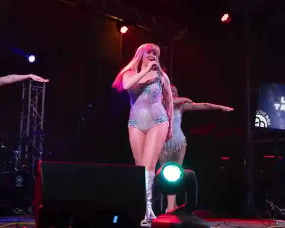 A performer in a sparkling silver outfit sings passionately on stage, with colorful lights illuminating the scene. Two backing dancers are positioned to the side, striking poses and enhancing the energetic atmosphere of the performance.